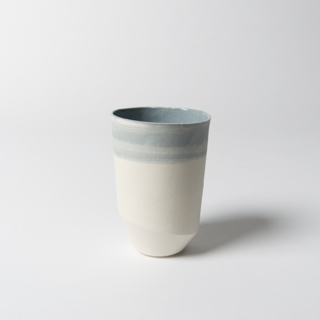 Foto zeigt eine Variante des asymmetrischen, schlanken Bechers aus weißem Porzellan mit Farbverläufen dank blauer Farbkörper