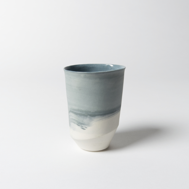 Foto zeigt eine Variante des asymmetrischen, schlanken Bechers aus weißem Porzellan mit Farbverläufen dank blauer Farbkörper