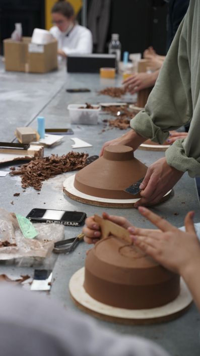 Foto zeigt Hände, die nach einem Claymodell einer Schale greifen und eine Modellierklinge halten. Unscharf im Vorder- und Hintergrund sind ebenfalls Hände und Claymodelle zu sehen