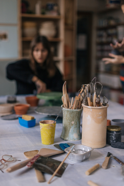 Foto zeigt Werkzeuge auf einem Tisch in einer Keramikwerkstatt stehen. Im Hintergrund sieht man eine Person Ton modellieren – ein Einblick in den Töpferkurs