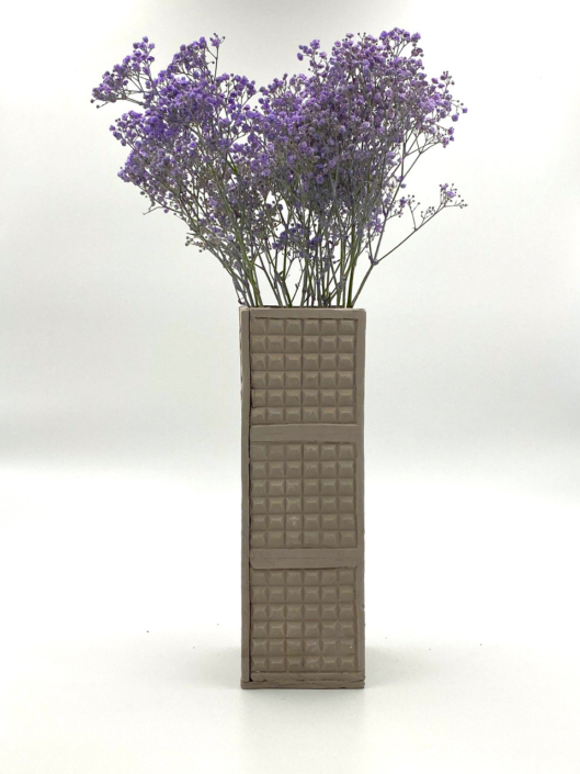 Bild zeigt rechteckige Vase mit einer Struktur, die an eine bestimmte Mauer in Russland erinnert. In der Vase sind lila Blumen.