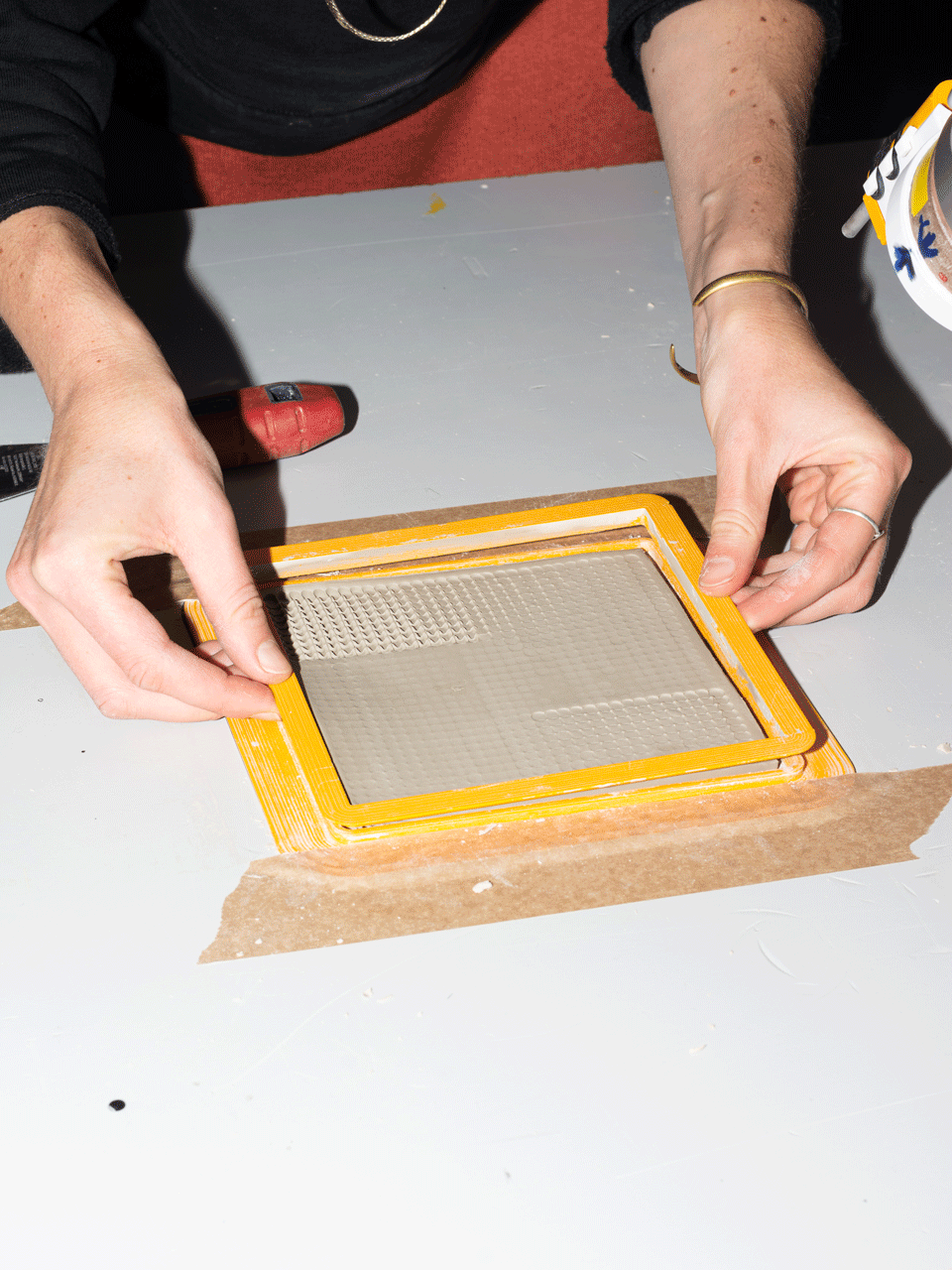 Hände greifen nach einem Rahmen, in dem eine strukturierte und noch feuchte Tonplatte liegt